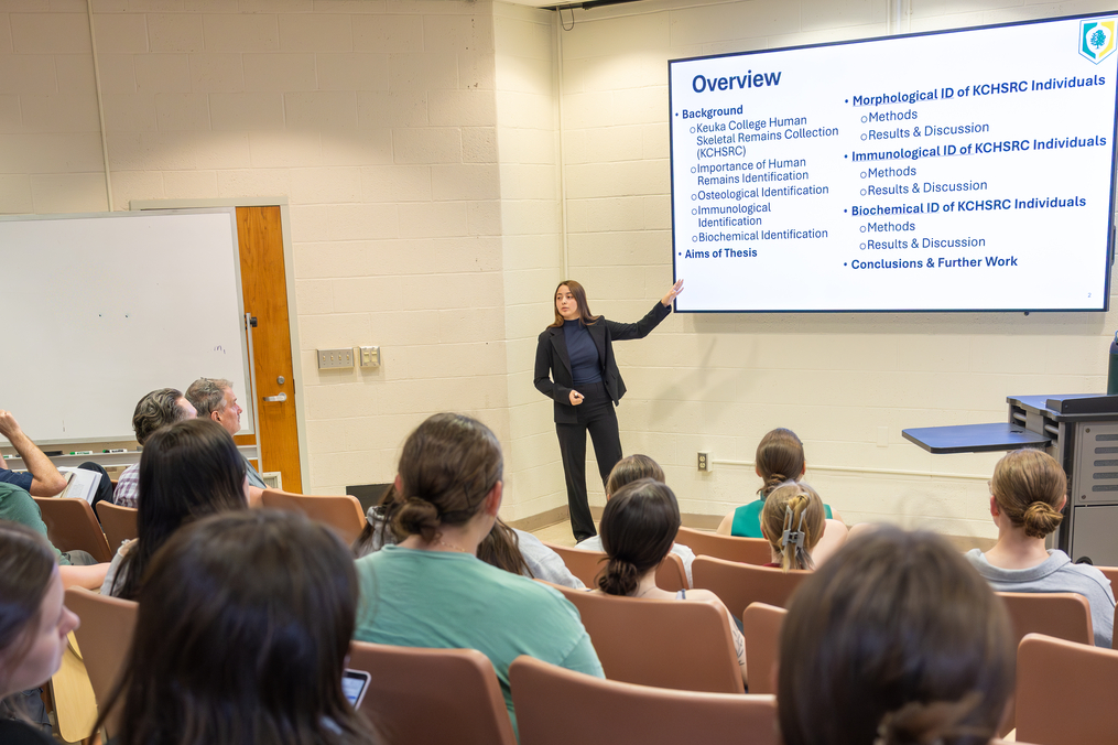 A student presenter stands at the front of a classroom, gesturing toward a large screen displaying a slide titled “Overview.” The slide outlines topics including background, methods, and conclusions related to human skeletal remains research. The presenter wears professional attire and faces an audience of seated students in a lecture hall, who are watching attentively. A whiteboard and classroom equipment are visible along the walls.