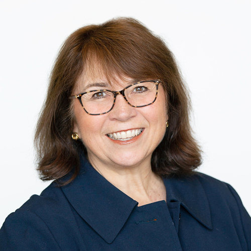 Professional headshot of a smiling woman with shoulder-length brown hair and glasses, wearing a navy-blue jacket and gold earrings against a light gray background. She faces the camera directly with a friendly, approachable expression.
